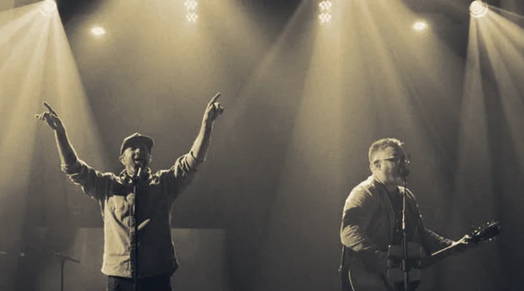 Two worship leaders on stage under spotlights, one with arms raised